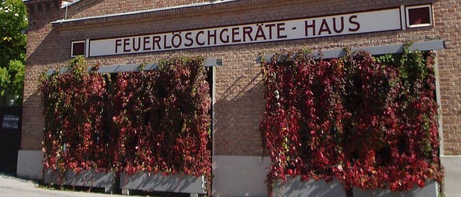 Ein Backsteingebäude mit einem Turm und einem Schild, das 'Feuerloschgerate-Haus' besagt, hat rote Reben, die die Vorderseite bedecken.