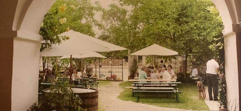 Ein Restaurant mit Sitzgelegenheiten im Freien unter Schirmen, mit Menschen, die an Tischen sitzen, einer Person, die mit einem Hund steht, und Topfpflanzen.