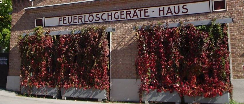 Ein Backsteingebäude mit einem weißen Turm trägt ein Schild mit der Aufschrift 'Feuerloschgerate Haus'. Das Gebäude ist mit roten Weinranken geschmückt, und links befindet sich ein Baum.