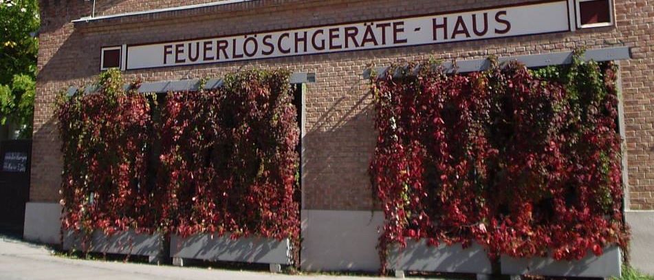 Ein Backsteingebäude mit einem weißen Turm, das das Schild 'Feuerlöschgeräte - Haus' zeigt. Rote Efeu wuchert an der Fassade.