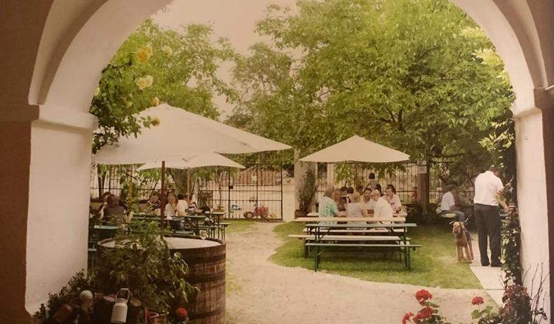Ein Vintage-Foto eines Gartenrestaurants mit Tischen, Stühlen und Schirmen. Menschen sitzen um die Tische. Eine Person steht in der Nähe des Tors. Bäume, Pflanzen und Blumen umgeben das Gebiet.