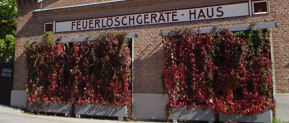 Ein Backsteingebäude mit einem Turmaufbau, das ein Schild mit der Aufschrift Feuerlöschgeräte-Haus zeigt. Weinreben bedecken die Vorderseite, mit einem Baum im Hintergrund.