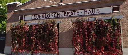 A brick building with a tower, named 'Feuerloschgerate-Haus', has a red vine on its front.