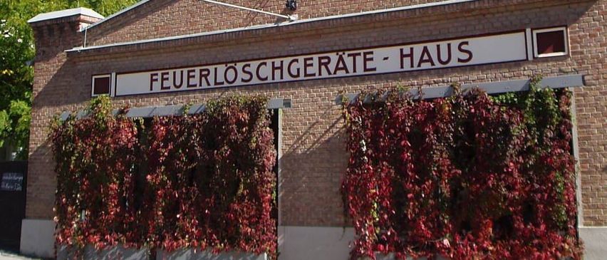 Ein Backsteingebäude mit einem Turm, benannt 'Feuerloschgerate-Haus', hat eine rote Rebe an der Vorderseite.