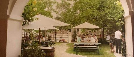 Eine Gruppe von Menschen sitzt unter weißen Sonnenschirmen an Tischen in einem Garten. Bäume und Pflanzen umgeben den Bereich. Ein Mann steht in der Nähe eines Hundes, und es gibt Fässer und Blumen in der Nähe.