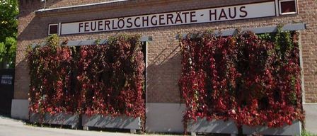 Ein Ziegelgebäude mit einem Turm und einem Schild, das 'Feuerloschgerate-Haus' steht, hat rote Kletterpflanzen an der Vorderseite.