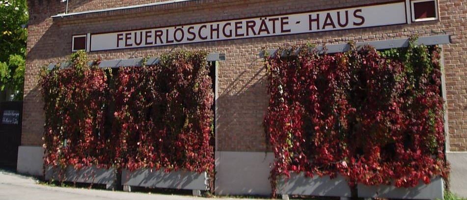 Ein Ziegelgebäude mit einem Turm und einem Schild, das 'Feuerloschgerate-Haus' steht, hat rote Kletterpflanzen an der Vorderseite.