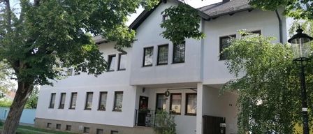 Ein weißes Haus mit vielen Fenstern, einem Schornstein und umgeben von Bäumen und Pflanzen. Ein Laternenpfahl steht auf der rechten Seite.