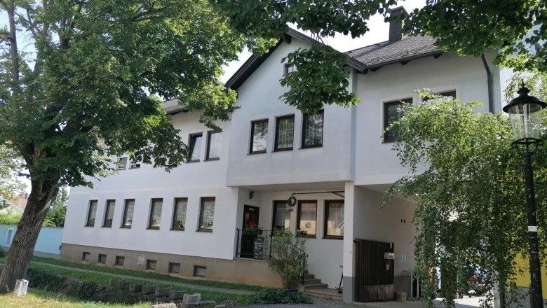 Ein großes weißes Gebäude mit mehreren Fenstern und einer schwarzen Tür, umgeben von üppiger Grünfläche und Bäumen.