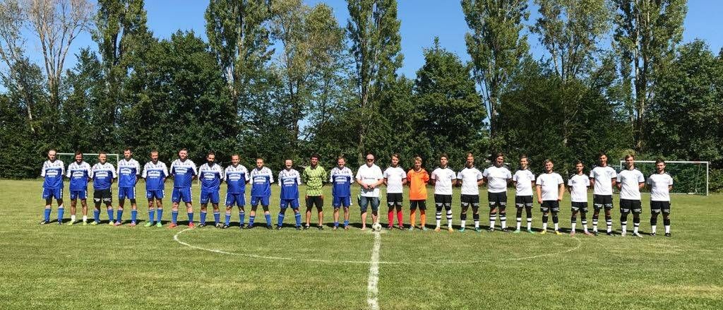 Eine Fußballmannschaft steht in einer Reihe auf einem Feld mit einer weißen Linie und grünem Gras. Die Spieler tragen verschiedenfarbige Trikots. Bäume und ein klarer blauer Himmel sind im Hintergrund.