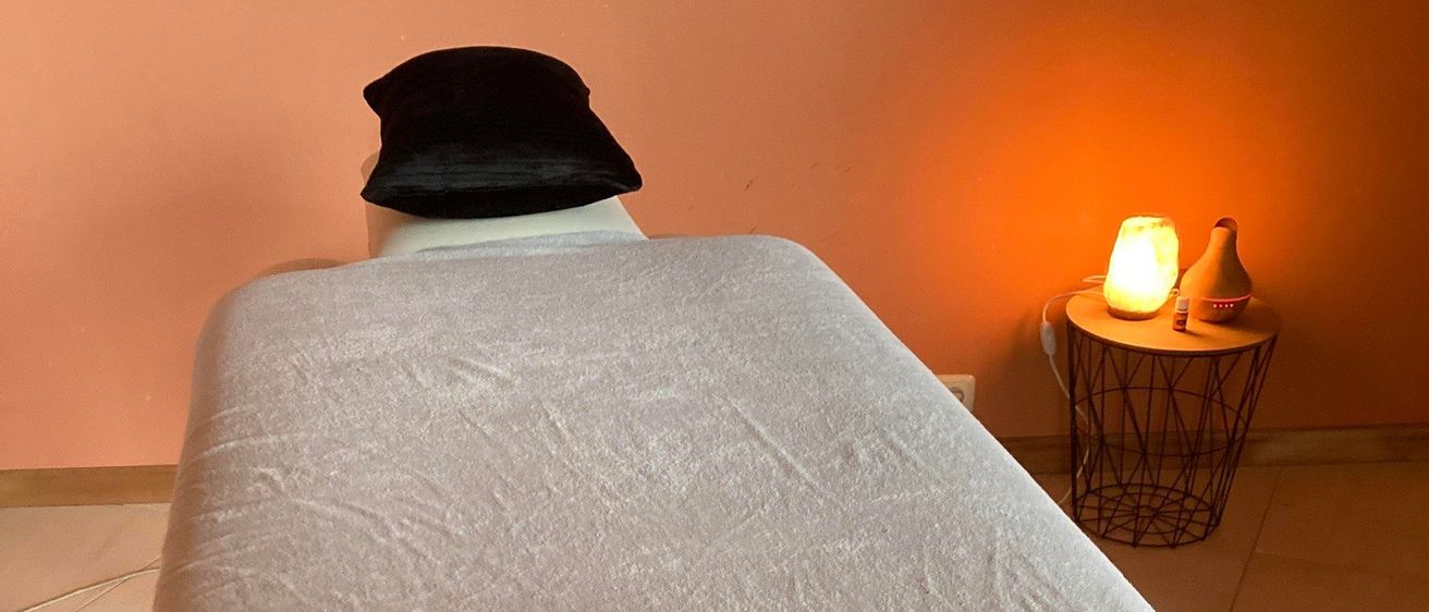 Ein Massagetisch mit Kissen und Handtuch, eine Lampe und eine Keramikvase auf einem Beistelltisch. Der Raum hat einen beigefarbenen Boden und pfirsichfarbene Wände.