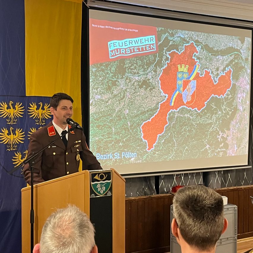A military officer stands at a podium speaking into a microphone. A large screen behind him displays a map with the words 'Feuerwehr Murstetten'.