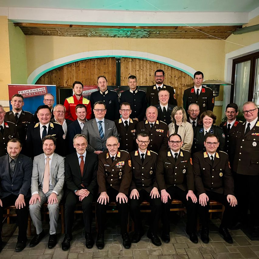 A group of uniformed men and women, possibly military officers, are posing for a photo in a room. Some are sitting while others are standing behind them.
