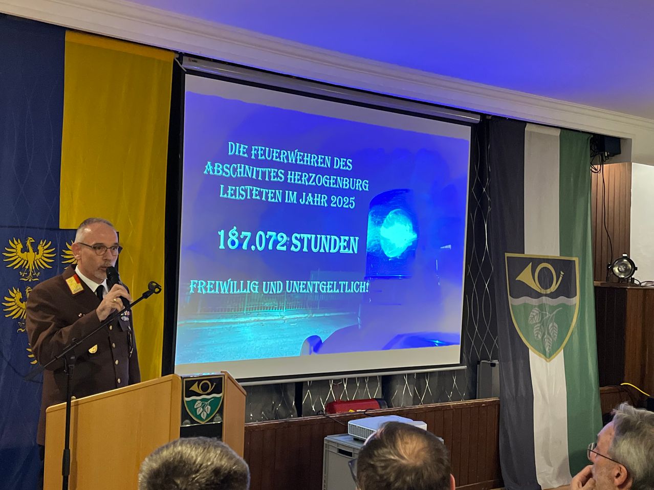 A man in uniform stands at a podium with a microphone, presenting information on a projector screen. The screen displays text in German, stating that Herzogenburg's firefighters volunteered 187,072 hours in 2025.
