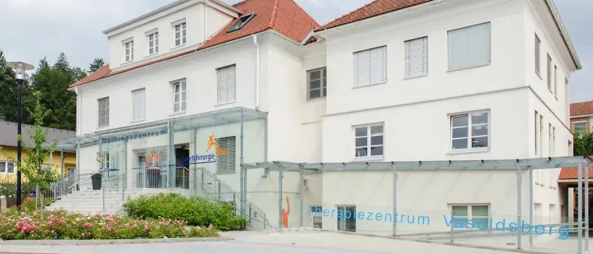 Ein modernes medizinisches Gebäude mit rotem Dach und weißen Wänden, mit einem gläsernen Eingang und mehreren Fenstern, steht vor einem Garten mit Blumen.