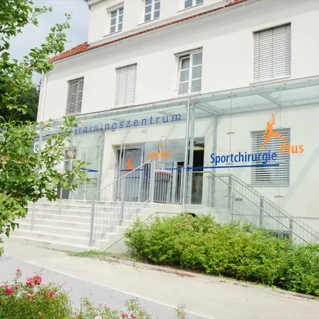Ein weißes Gebäude mit Glaseingang und Stufen. Es hat ein Schild, das 'Trainingszentrum' und 'Sportchirurgie Plus' besagt. Der Eingang hat ein Metallgeländer. Es gibt Pflanzen und Blumen vor dem Gebäude.