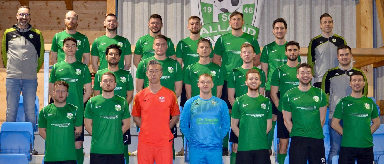 Eine Fußballmannschaft posiert für ein Gruppenfoto, wobei die Spieler grüne und orangefarbene Trikots tragen. Ein Banner mit der Nummer 19 und 46 steht hinter ihnen. Die Spieler stehen auf einer Plattform, wobei einige auf blauen Stühlen sitzen.