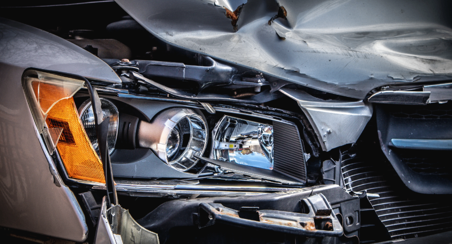 Nahaufnahme der durch eine Kollision beschädigten Fahrzeugfront. Der Stoßfänger und die Motorhaube sind verbogen, mit Kratzern und Dellen. Scheinwerfer sind beschädigt.