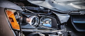 Nahaufnahme der durch eine Kollision beschädigten Fahrzeugfront. Der Stoßfänger und die Motorhaube sind verbogen, mit Kratzern und Dellen. Scheinwerfer sind beschädigt.