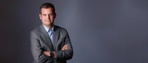 Ein Mann mit kurzen Haaren, der einen grauen Anzug, ein blaues Hemd und eine Uhr trägt, posiert mit verschränkten Armen für ein Foto.