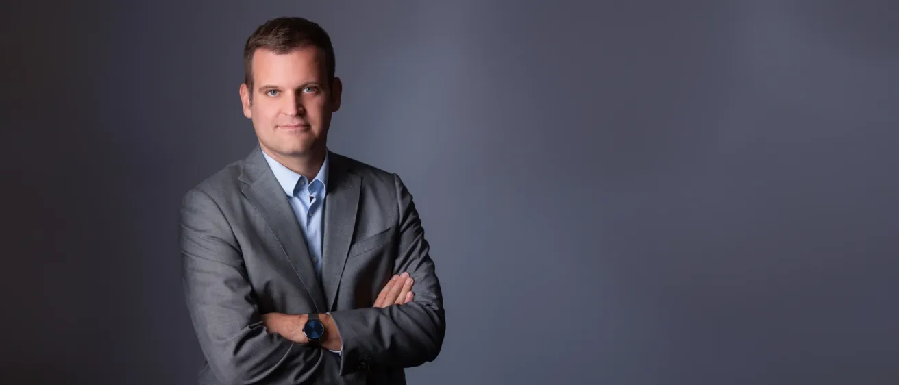 Ein Mann mit kurzen Haaren, der einen grauen Anzug, ein blaues Hemd und eine Uhr trägt, posiert mit verschränkten Armen für ein Foto.