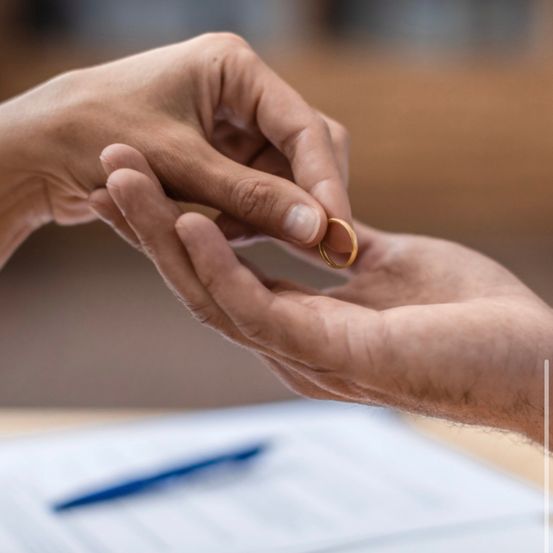 Ein Nahaufnahme von zwei Händen, die einen goldenen Ring halten, symbolisieren eine Verpflichtung oder Heiratsantrag. Der Hintergrund ist verschwommen und der Anlass ist feierlich.