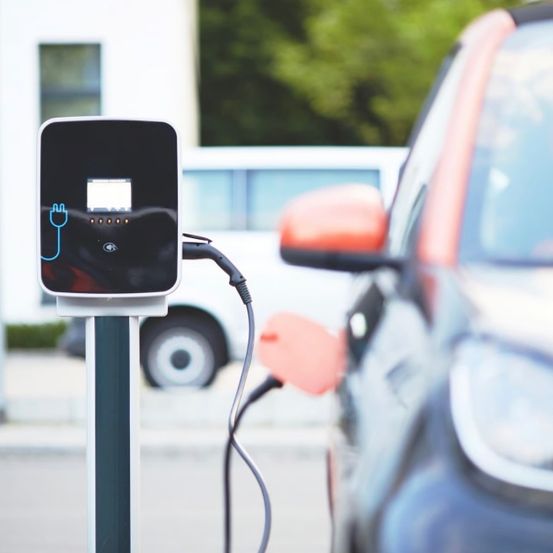 Ein Elektroauto wird an einer Ladestation aufgeladen. Ein weißer Lieferwagen ist dahinter geparkt. Die Szene spielt im Freien.