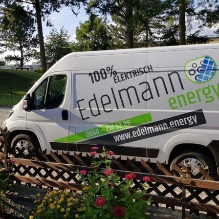 Ein weißer Lieferwagen mit grünen Akzenten ist vor einem Holzzaun geparkt. Auf der Seite des Lieferwagens steht '100% ELEKTRISCH' und 'Edelmann energy'. Es gibt ein Logo an der Seite des Lieferwagens.