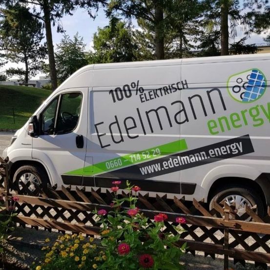 Ein weißer Lieferwagen mit grünen Akzenten ist vor einem Holzzaun geparkt. Auf der Seite des Lieferwagens steht '100% ELEKTRISCH' und 'Edelmann energy'. Es gibt ein Logo an der Seite des Lieferwagens.