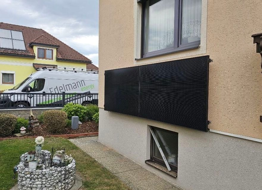 Ein Haus mit einem großen schwarzen Solarpanel an der Wand, einem geparkten Van davor und einem Garten mit einem Steinbrunnen im Vordergrund.
