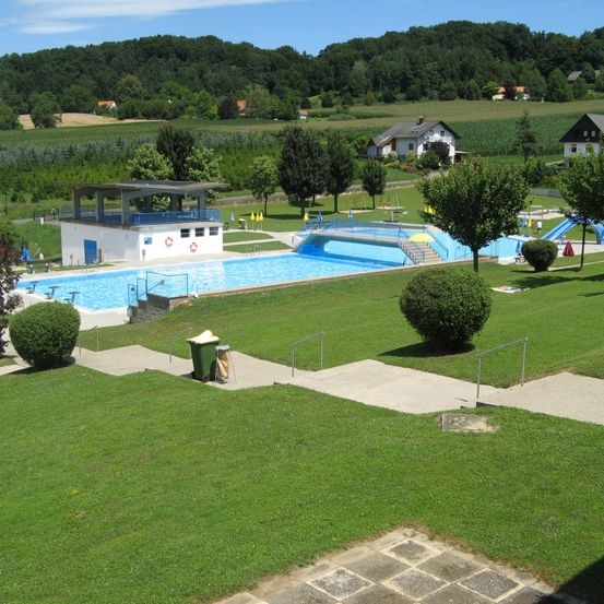 Ein Luftbild eines großen Freibades mit Sprungbrett und Rutsche, umgeben von gepflegten grünen Rasenflächen, Bäumen und Büschen. Der Pool wird von einem weißen Gebäude mit blauen Geländern gesäumt, und mehrere Häuser und ein Berg sind im Hintergrund zu sehen.