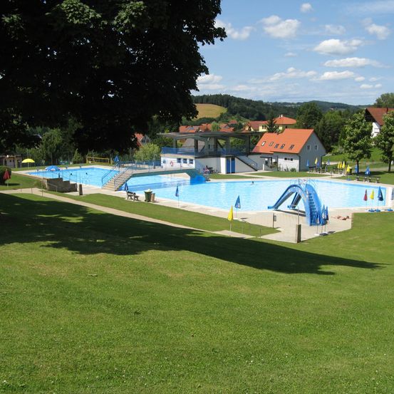 Ein Schwimmbereich mit Sprungbrett, mehreren Sonnenschirmen und klarem blauem Himmel. Im Hintergrund befinden sich Gebäude, Bäume und Berge.