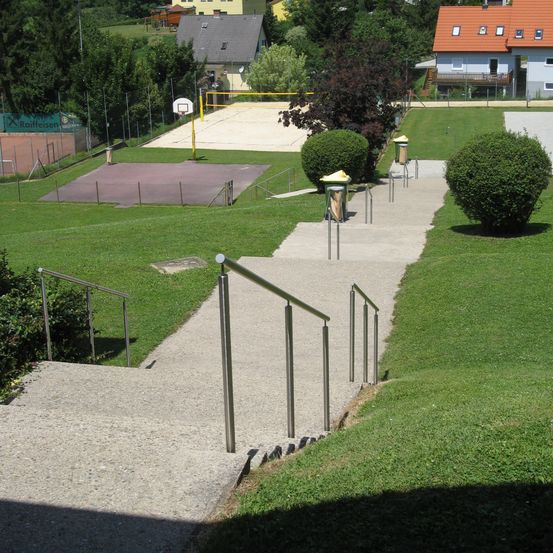 Ein Außenbereich mit einem Betonweg mit Metallhandläufen, der zu einem Spielplatz führt. Der Bereich umfasst einen Basketballplatz, ein Volleyballnetz und Häuser im Hintergrund.