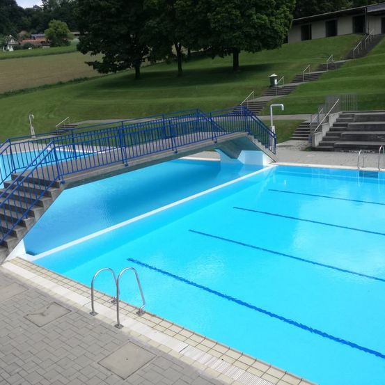 Ein Freibad mit einer blauen Brücke und Stufen darüber. Der Pool ist mit klarem blauem Wasser gefüllt und hat blaue Bahnen.