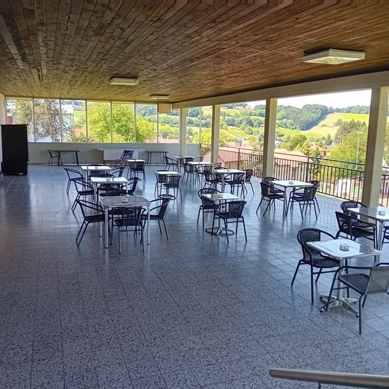 Eine leere Terrasse eines Restaurants mit gefliestem Boden und einer Holzdecke. Es gibt Tische und Stühle in Reihen angeordnet, und ein großes Glasfenster bietet einen Blick auf die Außenlandschaft.