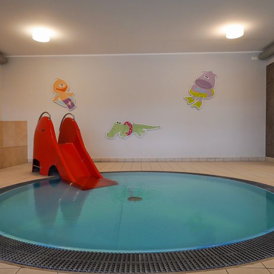 Ein kleiner Swimmingpool mit einer roten Rutsche, Cartoon-Tieren an der Wand und zwei Deckenleuchten.