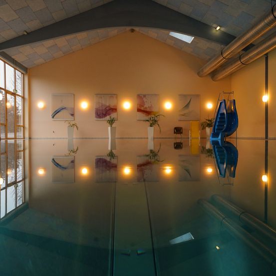 Ein Hallenbad mit einer Glasdecke, die Wände mit abstrakten Gemälden und Topfpflanzen reflektiert. Zwei blaue Rutschen werden auf dem Glas reflektiert.