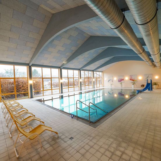 Ein Hallenbad mit blauem Wasser, einer blauen Rutsche und gelben Liegestühlen. Die Wände und die Decke sind weiß mit blauen Mustern, und es gibt große Fenster mit gelben Rahmen.