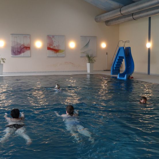 Ein Hallenbad mit drei Schwimmern und einer blauen Rutsche auf der rechten Seite. Der Pool hat einen blauen Boden und weiße Wände mit drei Gemälden.