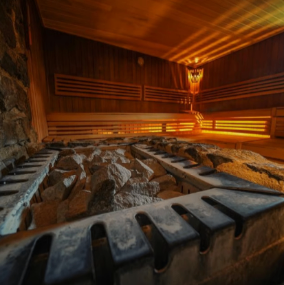 Ein gemütlicher Saunaraum mit Holzwänden, Bänken und einem Steinboden. Eine Lampe wirft ein warmes Licht in den Innenraum und hebt die Steine und Holzelemente hervor. Die Atmosphäre ist einladend und ruhig.
