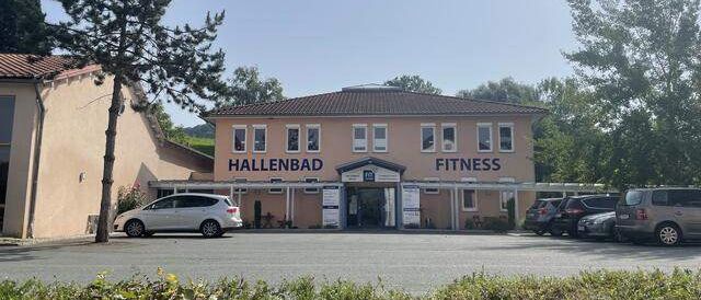Ein Außenfoto eines Fitnesscenters mit den Worten 'Hallenbad Fitness' auf der Vorderseite, mehrere Autos davor geparkt, ein Baum in der Mitte und Bäume in der Ferne.