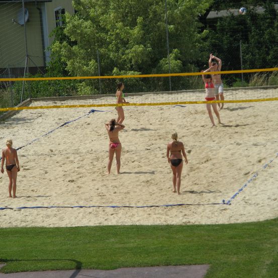 Eine Gruppe von Menschen spielt Volleyball auf einem Sandplatz. Ein Spieler hat gerade den Ball geschlagen.
