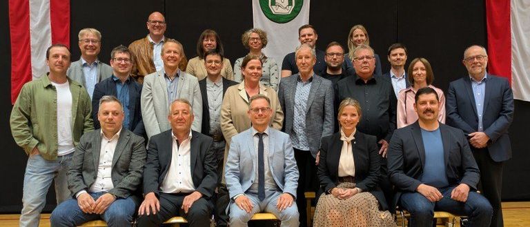 Eine Gruppe von Menschen in formeller Kleidung sitzt auf Stühlen vor einem schwarzen Hintergrund mit einer Flagge und einem Banner dahinter.