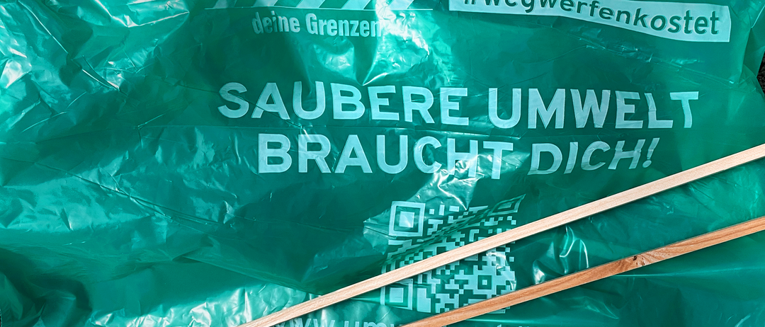 Ein grüner Plastikbeutel mit 'Respektiere deine Grenzen' und 'SAUBERE UMW BRAUCHT DICH'. Ein QR-Code und Holzstäbchen sind sichtbar.