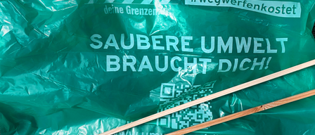 A green plastic bag with 'Respektiere deine Grenzen' and 'SAUBERE UMW BRAUCHT DICH'. A QR code and wooden sticks are visible.