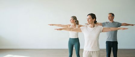 Zwei Frauen in einem Studio praktizieren Yoga, beide tragen weiße Shirts und Leggings. Eine in Türkis und die andere in Weiß.