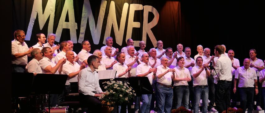 Ein Chor tritt auf der Bühne auf, mit einem Banner im Hintergrund, das Manner liest. Das Publikum klatscht, während die Musiker spielen.