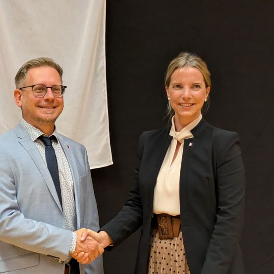 Ein Mann und eine Frau stehen und schütteln sich die Hände in einem Raum mit einer weißen Kulisse hinter ihnen.