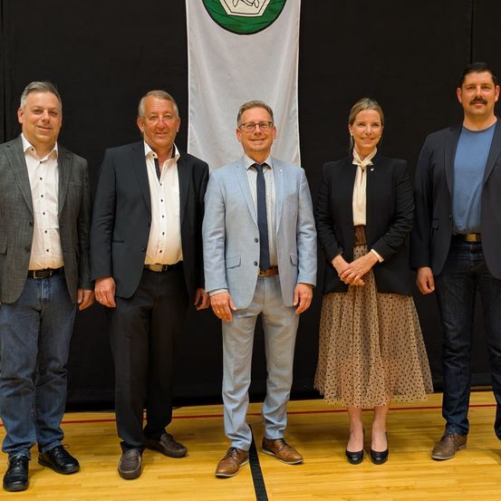 Fünf Personen in formeller Kleidung posieren für ein Foto in einem Raum. Sie stehen vor einer schwarzen Wand mit einem weißen Banner dahinter.