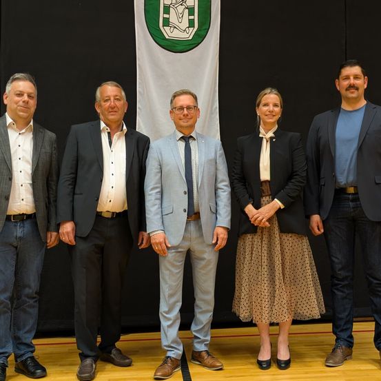 Fünf Erwachsene stehen in einer Reihe vor einer Flagge mit einem Logo. Die drei Männer tragen Anzüge und die beiden Frauen sind in Geschäftskleidung gekleidet.